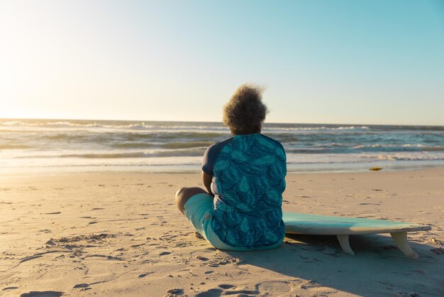 Rücksicht auf eine afrikanisch-amerikanische ältere Frau mit Surfbrett, die auf dem Strand sitzt und auf das Meer schaut.