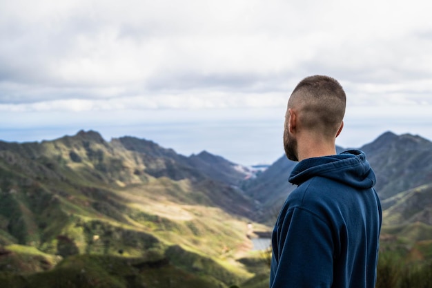 Rückwärts vom kaukasischen jungen Mann mit blauem Kapuzenpulli und bewölktem Tag, der die Landschaft betrachtet