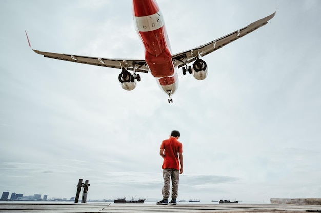 Rückwand des Jungen, der auf dem Pier steht, gegen ein Flugzeug, das im Himmel fliegt