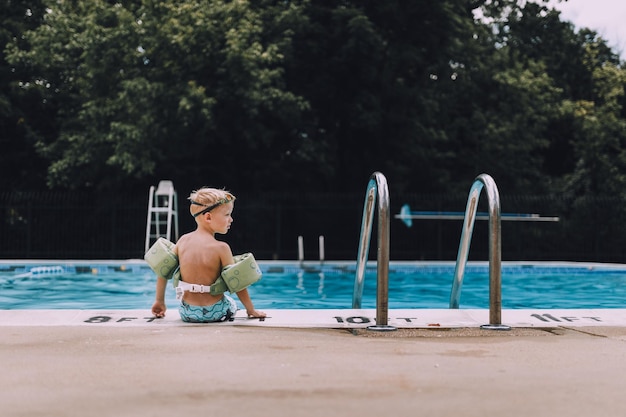 Rückwand eines hemdlosen Jungen mit Wasserflügeln, der am Pool sitzt