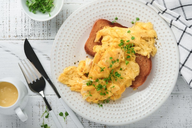 Rührei Fluffy and Buttery Rührei auf Brot mit mikrogrünem Rettich und Sauce Hollandaise auf weißem Teller auf weißem Holzhintergrund Hausgemachtes Frühstück oder Brunch Mahlzeit Draufsicht