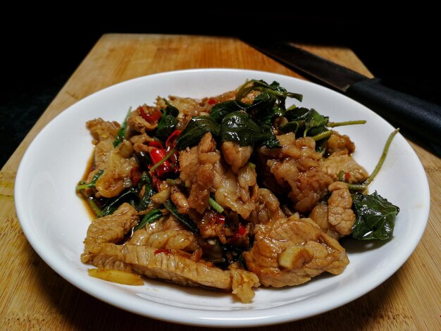 Rühren Sie gebratenes Rindfleisch mit heiligem Basilikum in einer weißen Platte, die auf einem Holzschneidbrett und einem Messer gelegt wird. Populäres Essen in Thailand besteht aus Fleisch, Knoblauch, frischen Chilischoten und Basilikumblättern.