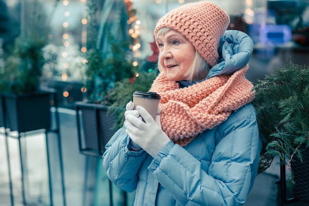 Ruhige gealterte Frau in Winterkleidung, die mit einer Tasse Kaffee in den Händen im Freien steht und in die Ferne schaut