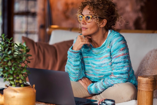 Ruhige hübsche erwachsene Frau zu Hause nach Online-Nutzung des Laptop-Computers, der auf dem Sofa sitzt und nach draußen schaut. Fröhliche moderne weibliche Menschen entspannen sich drinnen im Wohnzimmer und lächeln