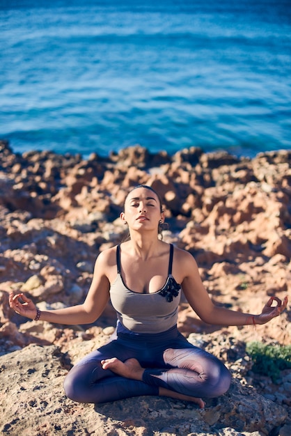 Ruhige junge Frau, die am Strand gegen ein schönes blaues Meer meditiert.