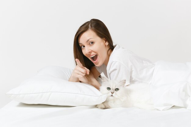 Ruhige junge Frau im Bett liegend mit weißer süßer persischer silberner Chinchilla-Katze, Laken, Kissen, Decke auf weißer Wand