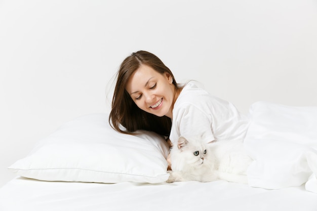 Ruhige junge Frau im Bett liegend mit weißer süßer persischer silberner Chinchilla-Katze, Laken, Kissen, Decke auf weißer Wand