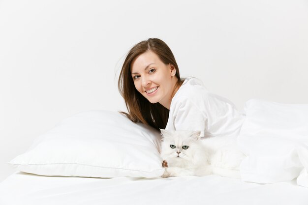 Ruhige junge Frau im Bett liegend mit weißer süßer persischer silberner Chinchilla-Katze, Laken, Kissen, Decke auf weißer Wand
