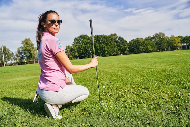 Ruhige junge Frau mit einem Golfschläger in der rechten Hand, die auf ihren Hintern sitzt