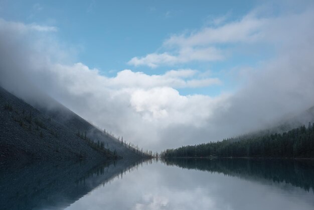 Ruhige, meditative, neblige Landschaft eines Gletschersees mit Spiegelung spitzer Tannenwipfel und Wolken am frühen Morgen. Grafischer EQ von Fichtensilhouetten am ruhigen Alpenseehorizont. Bergsee im Nebel