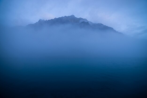 Ruhige, minimale Landschaft mit großem felsigen Berg mitten im Wasser zwischen niedrigen Wolken in der Dämmerung. Wellenförmiges Meer der blauen klassischen Farbe. Atmosphärische Landschaft mit tiefblauem, ruhigem See und Felsen im dichten Nebel.