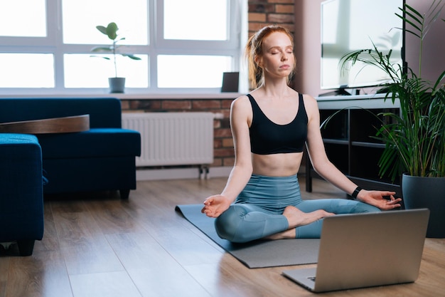 Ruhige rothaarige junge Frau mit geschlossenen Augen, die zu Hause meditiert und in Lotusposition auf einer Yogamatte in der Nähe eines Laptop-Computers sitzt Friedliche rothaarige Dame, die die Hände auf dem Knie in OM-Position hält