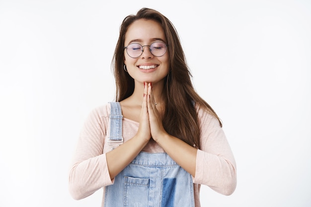 Ruhige und friedliche, glückliche buddhistische Frau in Brille und Overall, die Handflächen über der Brust zusammenhält, wie beim Beten und mit geschlossenen Augen lächelt, Namaste sagt und sich freundlich über graue Wand verbeugt.