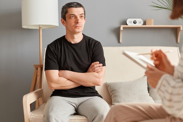 Ruhiger, gleichgültiger kaukasischer Mann, der auf der Couch in der Psychologiepraxis sitzt und einer Therapeutin zuhört, die in einer Therapiesitzung psychische Probleme löst