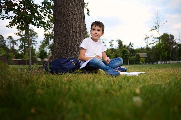 Ruhiger hübscher Schuljunge, der Hausaufgaben macht, auf grünem Gras des Stadtparks sitzt, zur Seite schaut und während seiner Erholung nach dem ersten Schultag frische Luft genießt. Zurück zum Schulkonzept. September.