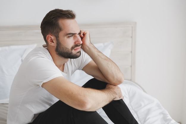 Ruhiger junger gutaussehender bärtiger Mann, der im Bett mit weißer Bettdecke im Schlafzimmer zu Hause sitzt