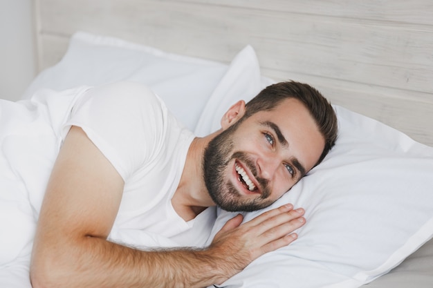 Ruhiger junger gutaussehender bärtiger Mann, der zu Hause mit weißer Kissendecke im Schlafzimmer im Bett liegt lying