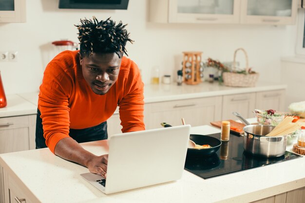 Ruhiger junger Mann, der aufmerksam auf den Bildschirm des modernen Laptops schaut, während er in der Küche mit der Pfanne an seiner Seite ist