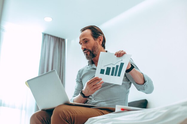 Ruhiger junger Mann, der der Laptop-Webkamera ein grafisches Diagramm zeigt, während er zu Hause auf dem Bett sitzt