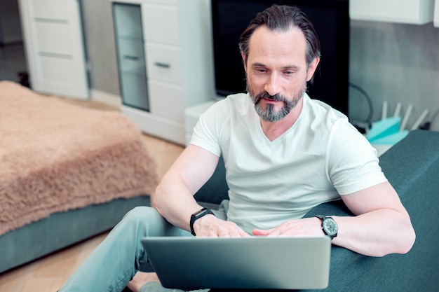 Ruhiger kaukasischer Mann, der allein im Schlafzimmer sitzt und auf den Laptopbildschirm schaut