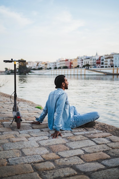 Ruhiger Mann, der mit gekreuzten Beinen neben dem Fluss in der Stadt sitzt. Er ist in der Nähe eines Elektrorollers