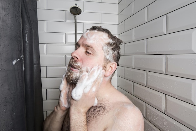 Ruhiger, nachdenklicher Mann schäumt sich mit Waschmittel ein, das in der Dusche unter einem Wasserstrahl steht