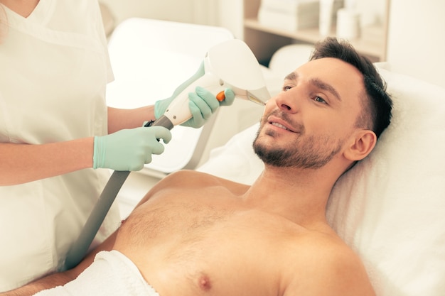 Ruhiger, selbstbewusster Mann, der sich dem Laser-Haarentfernungsverfahren mit einem modernen Werkzeug in der Nähe seines Gesichts unterzieht