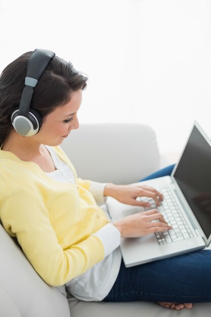 Ruhiger zufälliger Brunette in der gelben Wolljacke hörend Musik bei der Anwendung eines Laptops