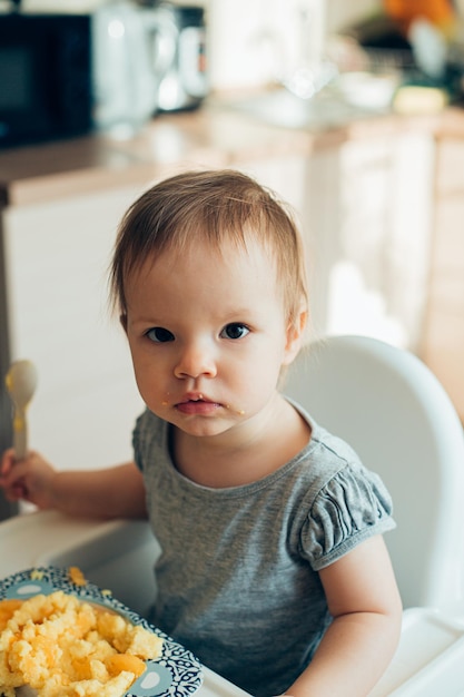 Ruhiges, neugieriges Kleinkind, das die Kamera anschaut, während es auf einem Kinderhochstuhl mit einem Teller Haferbrei sitzt