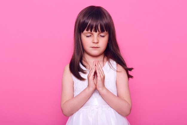 Ruhiges schönes kleines Mädchen im weißen Kleid, das Yoga praktiziert, Augen geschlossen hält, Handflächen zusammenpresst, dunkelhaariges süßes Kind betet, Hoffnung ausdrückt, isoliert über rosa Wand