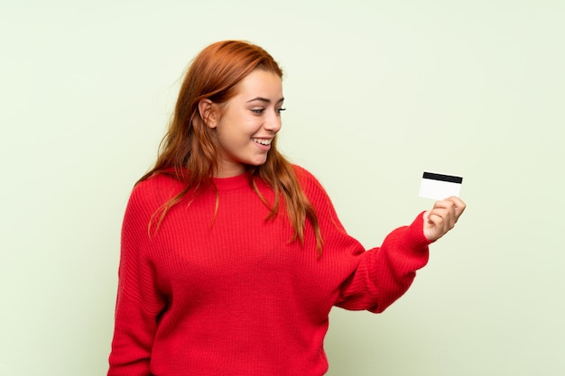 Ruiva adolescente com camisola sobre fundo verde isolado, segurando um cartão de crédito