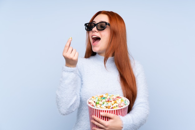 Ruiva adolescente menina isolada parede azul com óculos 3d e segurando um grande balde de pipocas