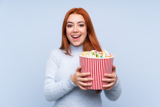 Ruiva adolescente menina isolada parede azul segurando um grande balde de pipocas