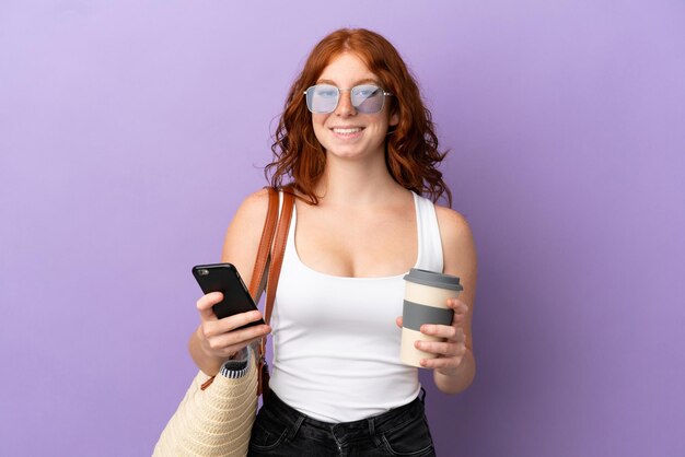 Ruiva adolescente segurando uma bolsa de praia isolada no fundo roxo segurando um café para levar e um celular