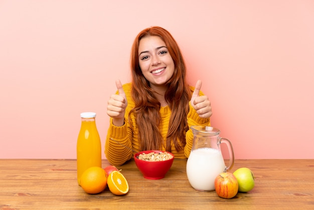 Ruiva adolescente tomando café da manhã em uma mesa dando um polegar para cima gesto