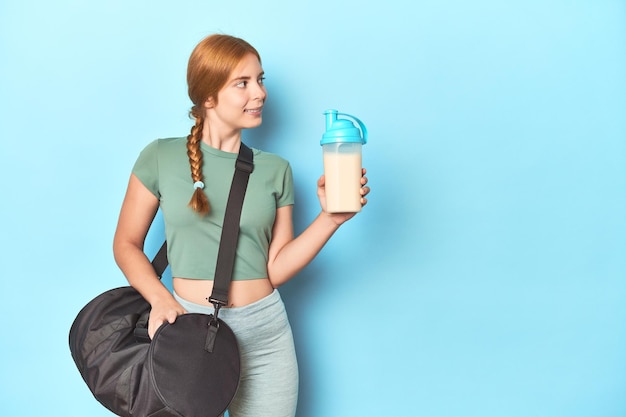 Ruiva atlética segurando shake de proteína e garrafa no estúdio azul