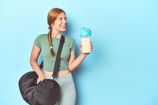 Ruiva atlética segurando shake de proteína e garrafa no estúdio azul