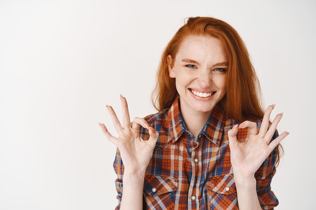 Ruiva atrevida mostrando sinais de ok e sorrindo feliz, recomendando produto, dizendo sim à boa escolha, elogiando algo, parede branca