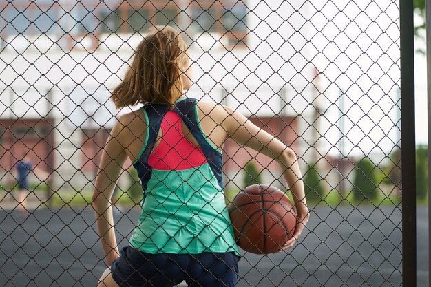 Ruiva caucasiana magra em pé perto da cerca da quadra de basquete de rua ao ar livre, segurando uma bola e observar um jogo