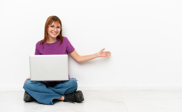 Ruiva com um laptop sentado no chão, estendendo as mãos para o lado para convidar para vir