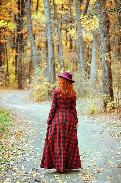 Ruiva de vestido vermelho, chapéu andando na floresta de outono. estilo de vida feliz. Outono. Folhas amarelas