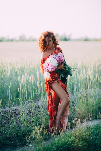 Ruiva de vestido vermelho com buquê de peônias dançando alegre no campo de trigo no verão ao pôr do sol. Vertical