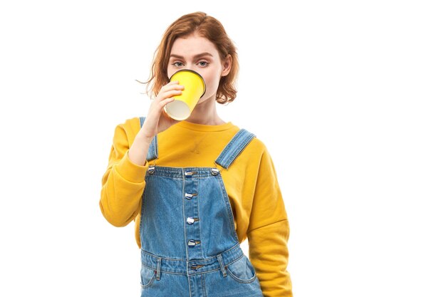 Ruiva enérgica segurando e bebendo uma xícara amarela de café para viagem sorrindo e desfrutando de uma bebida isolada no fundo branco