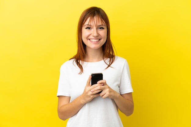 Ruiva isolada em fundo amarelo, olhando para a câmera e sorrindo enquanto estiver usando o celular