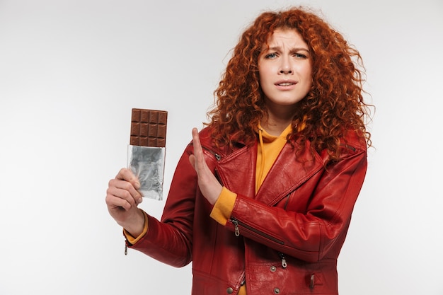 Foto ruiva saudável de 20 anos vestindo uma jaqueta de couro segurando uma barra de chocolate e expressando antipatia isolada sobre uma parede branca