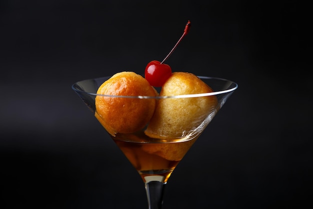 Rum napolitano baba ou baba au rum em um copo de martini com uma cereja de coquetel em um fundo preto Pequenos bolos de fermento embebidos em calda de rum Pastelaria italiana tradicional Closeup foco seletivo