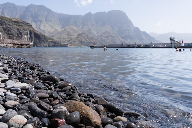 Runde Kieselsteine am Strand von Agaete mit Menschen auf dem Wasser am Pier und grünen Bergklippen