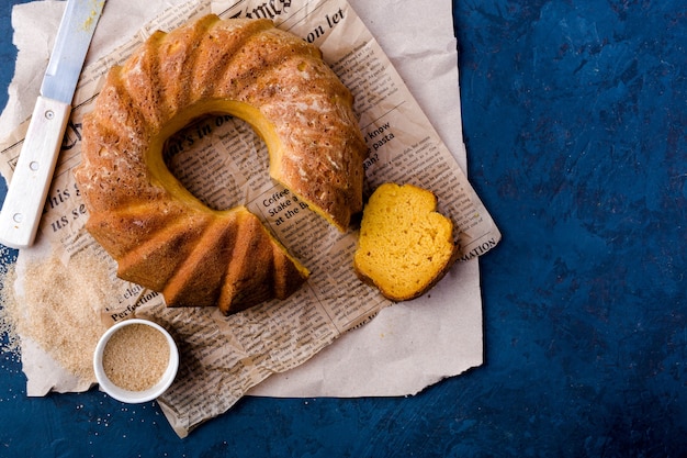 Foto runder kürbiskuchen, auf kraftpapier, messer, sauciere, brauner zucker, stück kuchen, draufsicht, dunkelblauer hintergrund