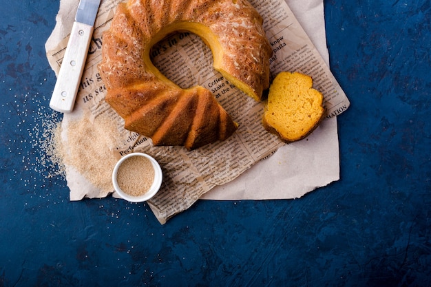 Foto runder kürbiskuchen, auf kraftpapier, messer, sauciere, brauner zucker, stück kuchen, draufsicht, dunkelblauer hintergrund