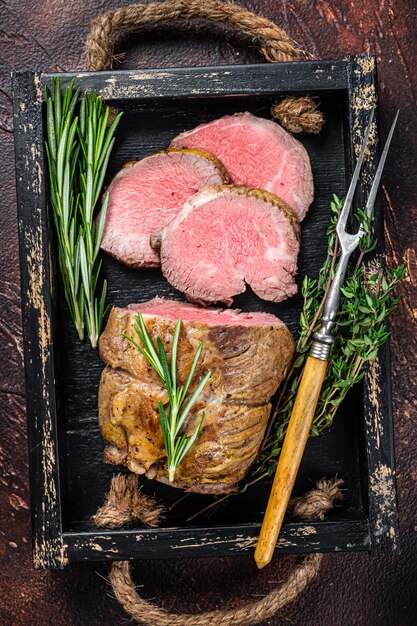 Rundes Filetfleisch vom Roastbeef in einem Holztablett mit Kräutern. Dunkel